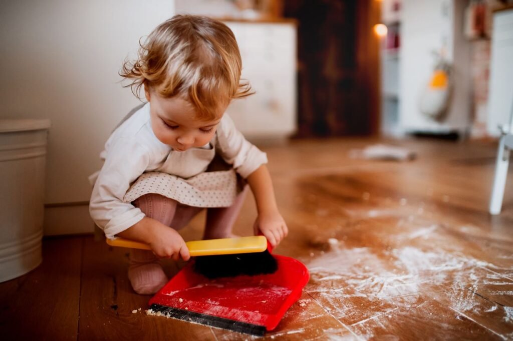 A toddler emulates household chores, a sign of early learning and responsibility, by tidily sweeping up a flour spill on the hardwood flooring of a well-maintained residential kitchen, indicating a family-friendly and lived-in aspect of the home.