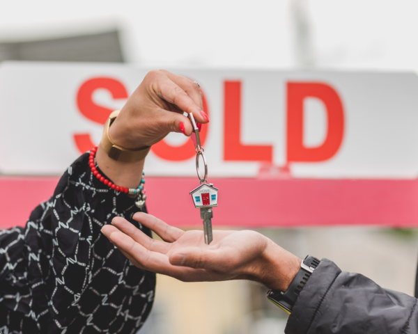 A close-up of two hands in front of a 'Sold' real estate sign, one hand passing a set of keys with a house-shaped keychain to the other, symbolizing the completion of a property sale and give an offer to homeowners