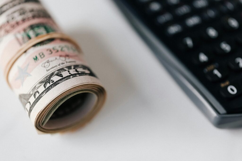 A roll of U.S. dollar bills secured with a rubber band, suggesting financial planning or savings, lies in focus beside a blurred calculator, symbolizing budgeting, investments, or accounting tasks.