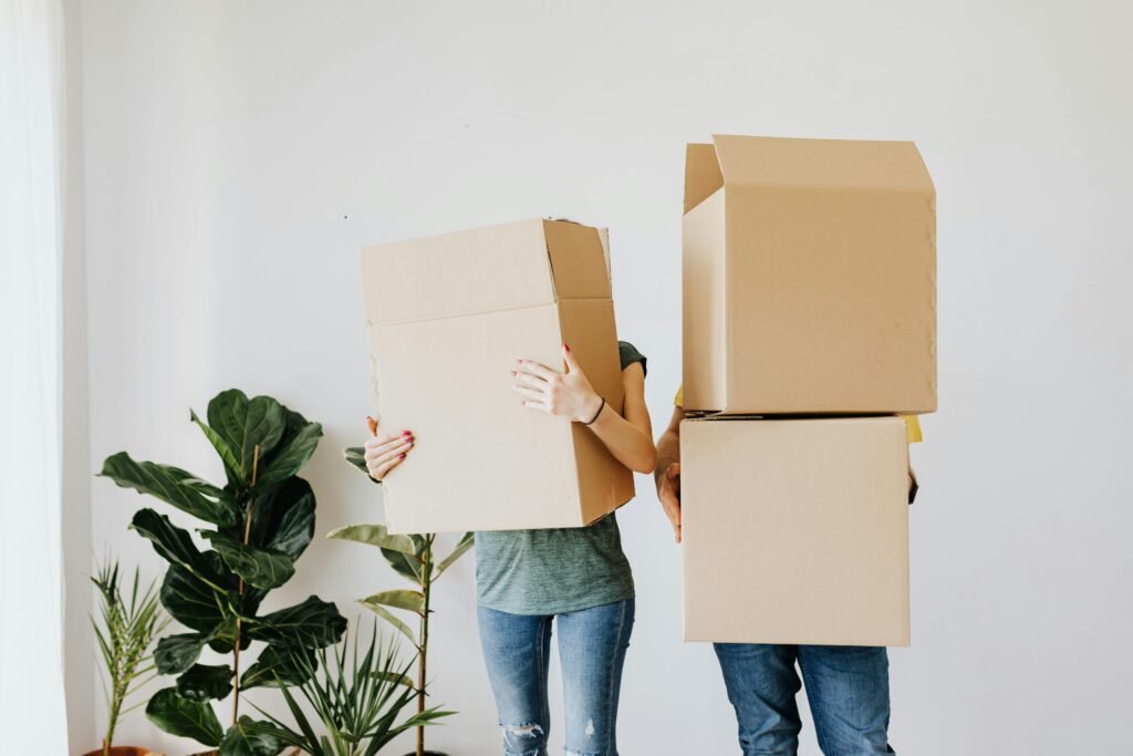 Two people standing in front of a white wall, each holding a large cardboard box in front of their upper bodies, obscuring their heads from view. The person on the left appears to be wearing a gray top and blue jeans with a visible pink manicure, while the person on the right is in a yellow shirt and blue jeans. In the background, there's an assortment of potted plants, including a large fiddle-leaf fig, adding a touch of greenery to the scene.