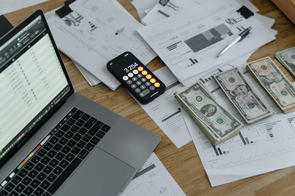 A busy financial workspace featuring an open laptop with a spreadsheet displayed on the screen, a smartphone showing a calculator app with the number 3,254 on the screen, and stacks of US dollar bills arranged next to a pen. There are also multiple papers with financial charts, graphs, and text scattered across the wooden surface, suggesting a thorough review or analysis of financial data is in progress.